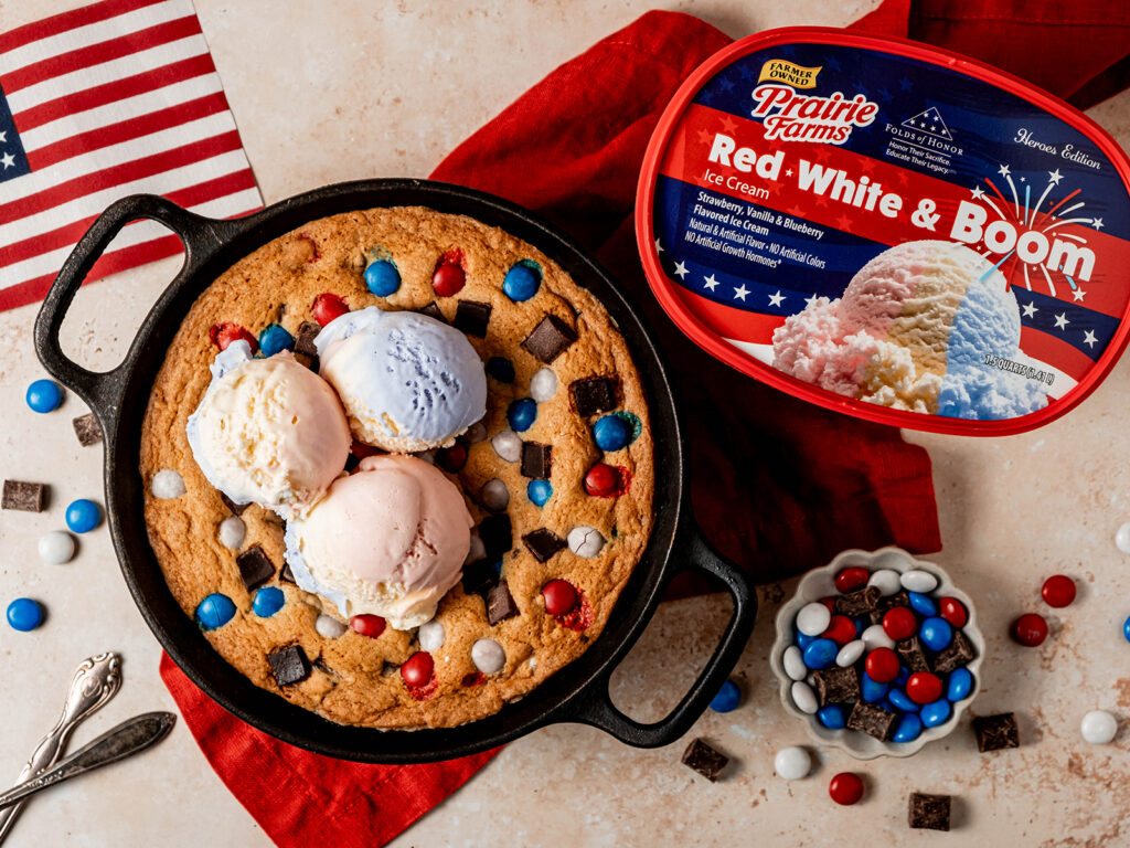 A patriotic skillet cookie topped with scoops of red, white, and blue ice cream, surrounded by colorful candy pieces and chocolate chunks, served in a cast iron skillet beside a container of Prairie Farms Red, White & Boom ice cream.
