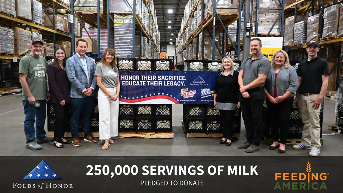 Group of representatives from Folds of Honor, Prairie Farms, Hiland Dairy, and Feeding America standing in a warehouse beside pallets of milk crates and a banner announcing a pledge to donate 250,000 servings of milk to support food banks and military families.