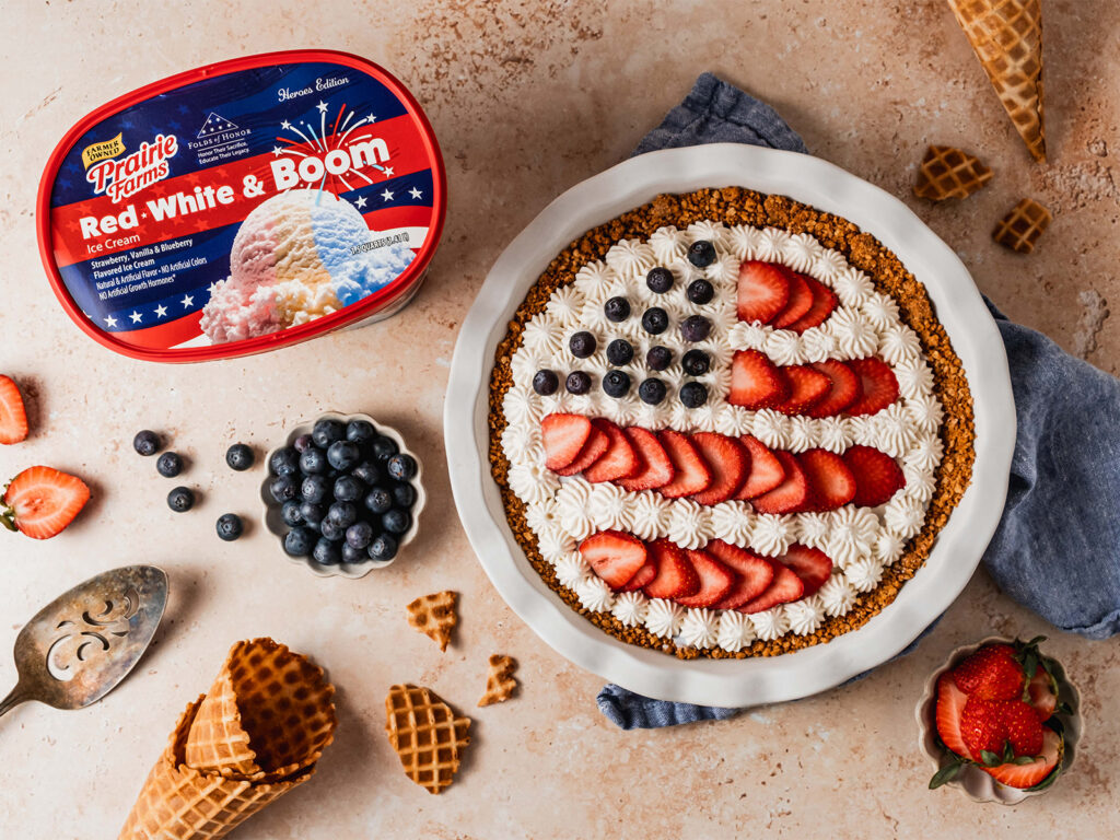 A patriotic ice cream pie decorated with whipped cream, strawberries, and blueberries in an American flag design, served in a waffle cone crust beside a container of Prairie Farms Red, White & Boom Ice Cream.