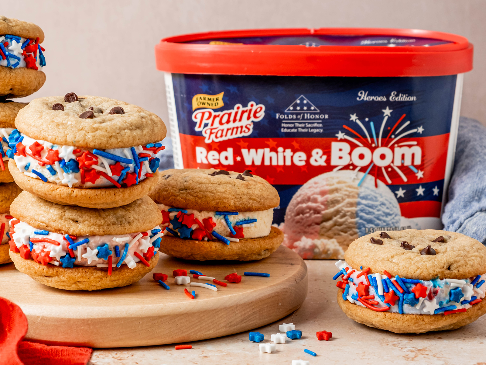 Chocolate chip cookie ice cream sandwiches filled with red, white, and blue ice cream and coated in patriotic sprinkles, arranged on a wooden board beside a container of Prairie Farms Red, White & Boom Ice Cream.
