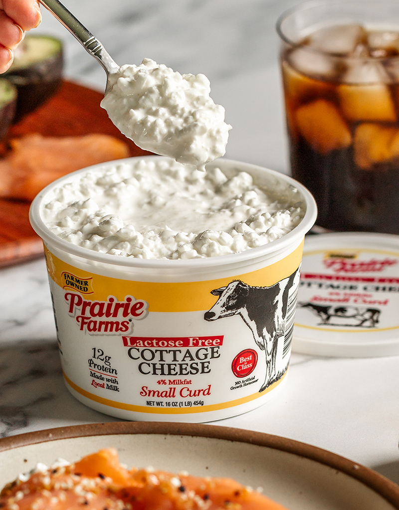 Spoon lifting Prairie Farms Lactose Free Small Curd Cottage Cheese from an open container with smoked salmon and iced coffee in the background