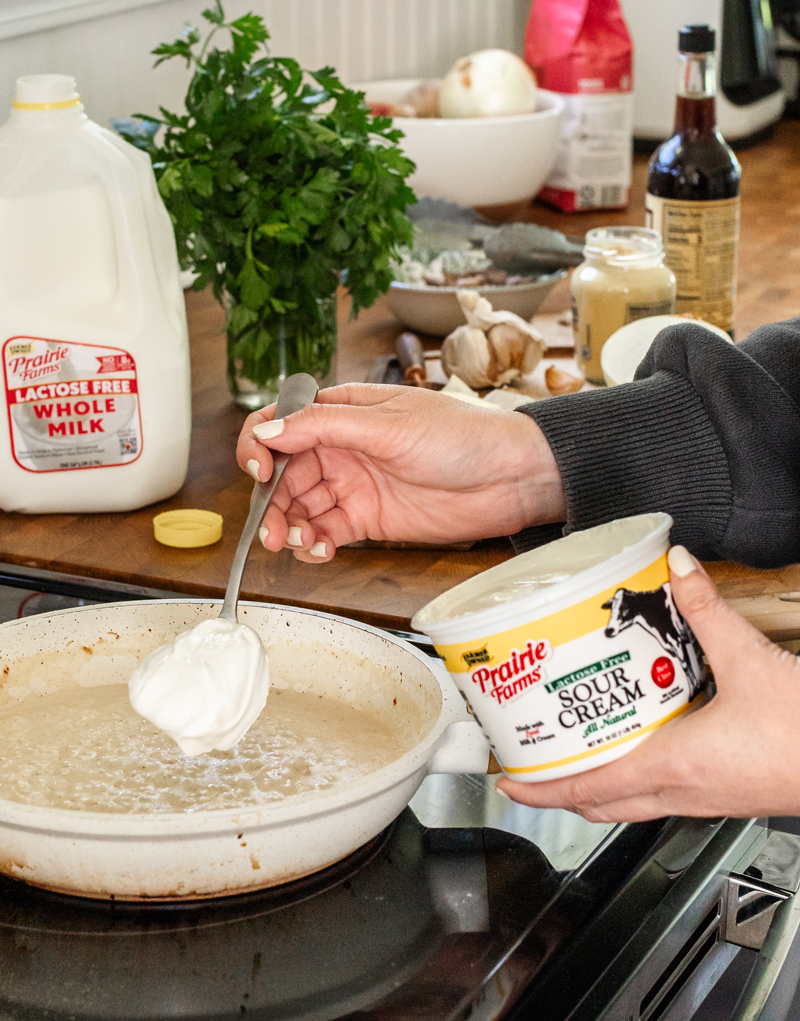 Prairie Farms Lactose Free Sour Cream held beside a skillet while a spoonful is added to a creamy sauce, with Prairie Farms Lactose Free Whole Milk and fresh cooking ingredients in the background