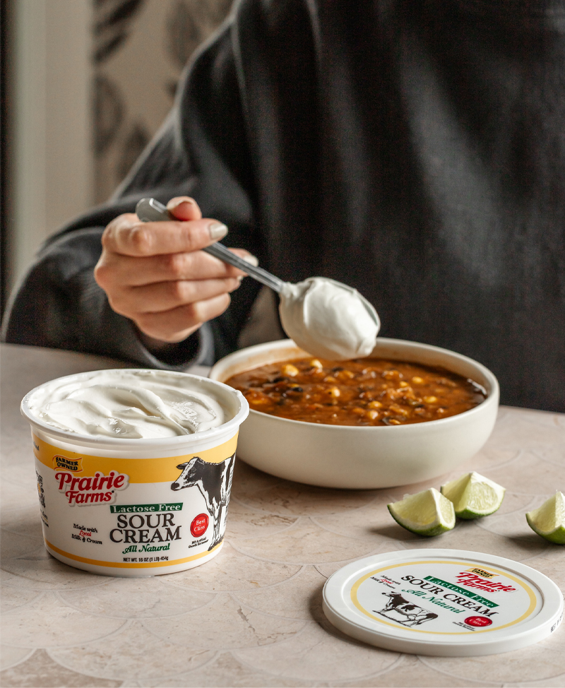 Prairie Farms Lactose Free Sour Cream beside a bowl of chili as a spoonful of sour cream is added, with fresh lime wedges on the table.