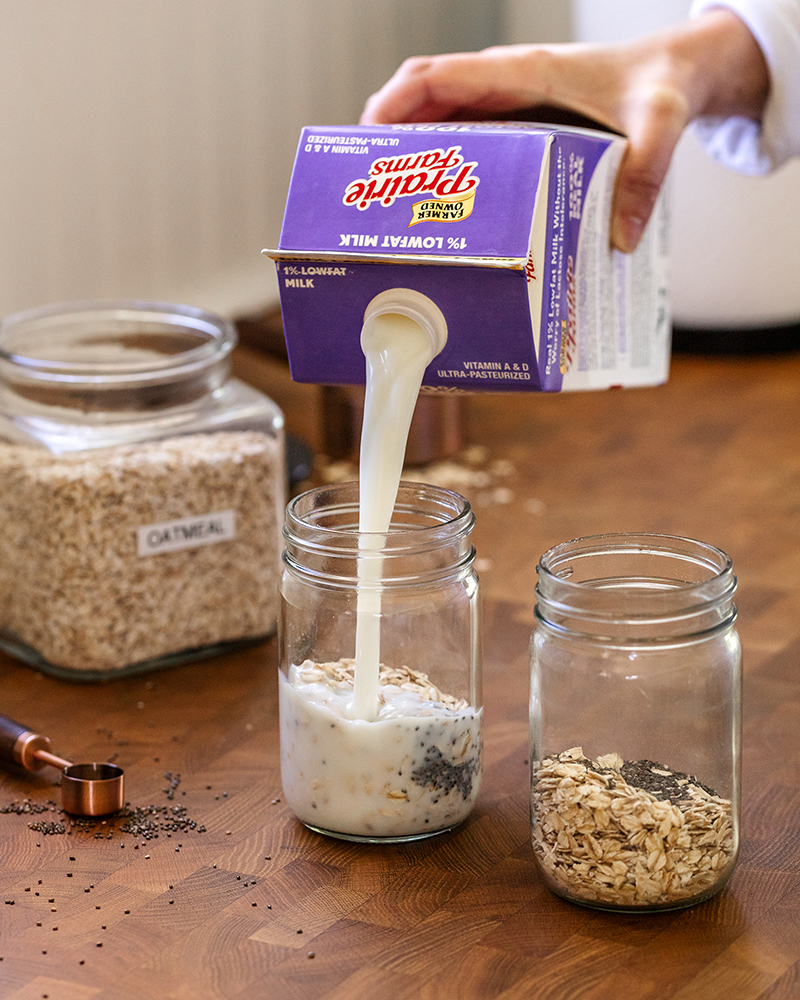 Prairie Farms 100% Lactose Free 1% Lowfat Milk being poured into a mason jar of overnight oats on a kitchen countertop.