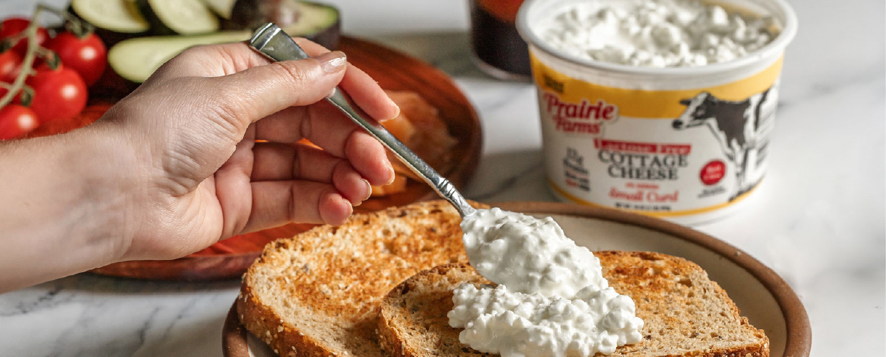 A hand spreading Prairie Farms Lactose Free Cottage Cheese onto toasted bread with fresh vegetables and an open cottage cheese container in the background