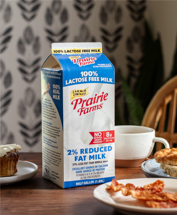 Prairie Farms 100% Lactose Free 2% Reduced Fat Milk half-gallon carton on a breakfast table with bacon, toast, and coffee