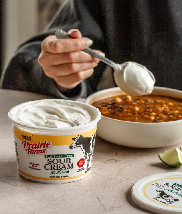 A spoonful of Prairie Farms Lactose Free Sour Cream being added to a bowl of chili with the sour cream container in the foreground