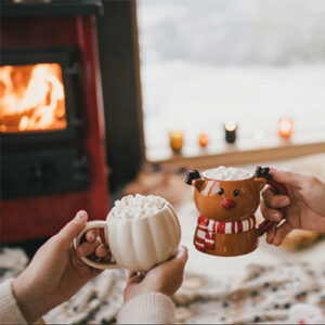 Two people holding festive mugs topped with marshmallows beside a cozy fireplace.