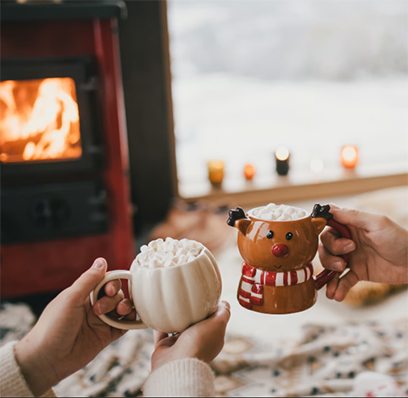 Two people holding festive mugs topped with marshmallows beside a cozy fireplace.