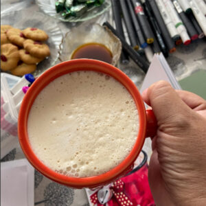 Red mug filled with a creamy holiday drink on a table with art supplies and cookies.