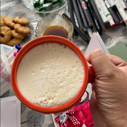 Red mug filled with a creamy holiday drink on a table with art supplies and cookies.