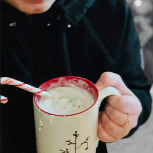 Person holding a holiday mug with a candy cane while enjoying a warm drink outdoors.