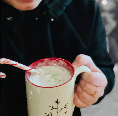 Person holding a holiday mug with a candy cane while enjoying a warm drink outdoors.