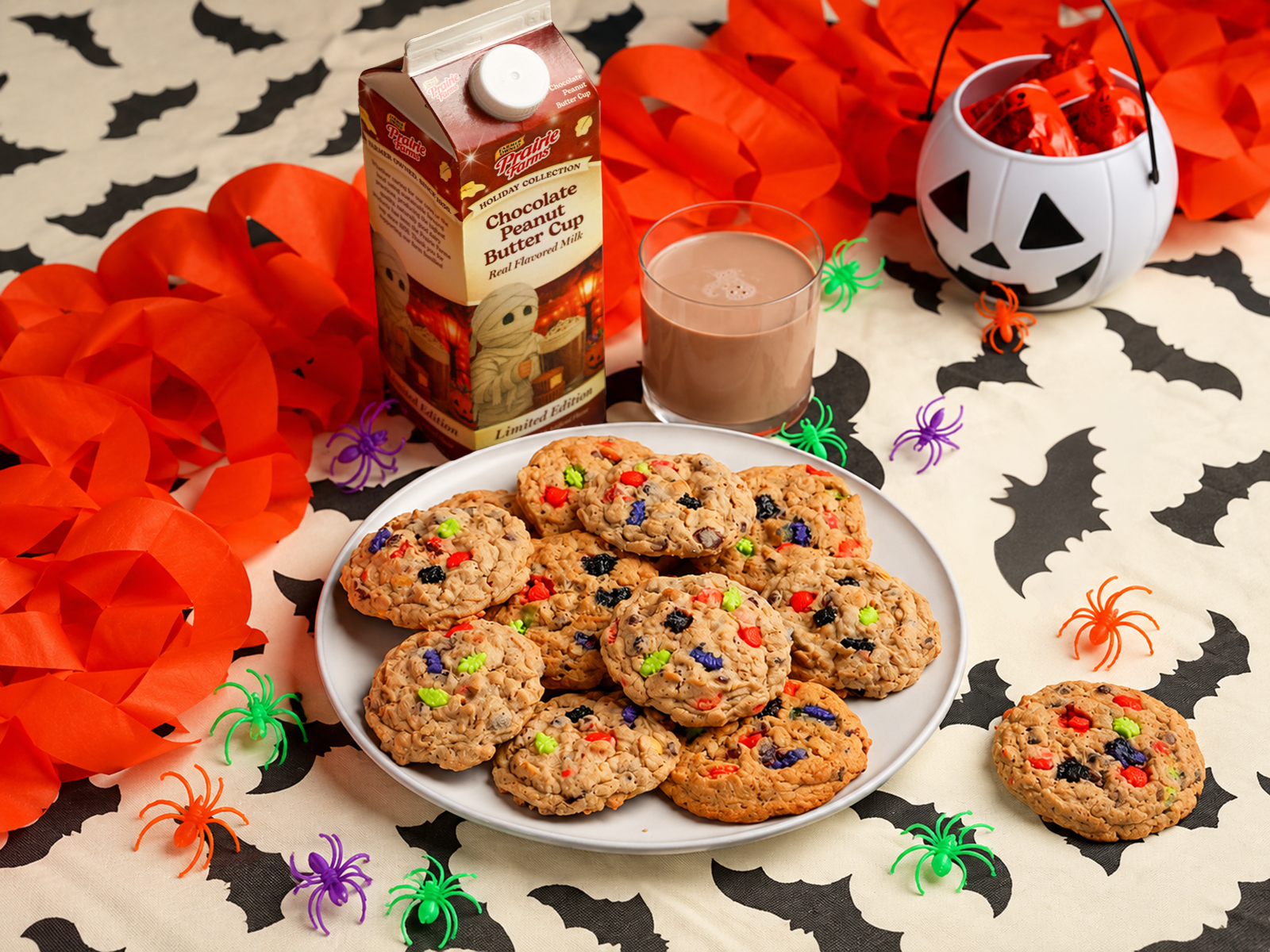 Chocolate peanut butter monster cookies filled with chocolate chips and colorful Halloween sprinkles, served with a glass of Prairie Farms Chocolate Peanut Butter Cup Milk among festive bats and spiders.