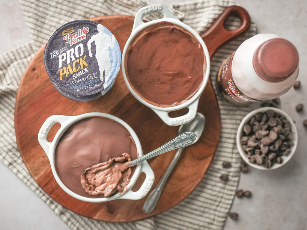 Overhead view of two chocolate protein cheesecake cups in small baking dishes on top of a circular wooden serving board, with one cracked open showing the creamy texture.