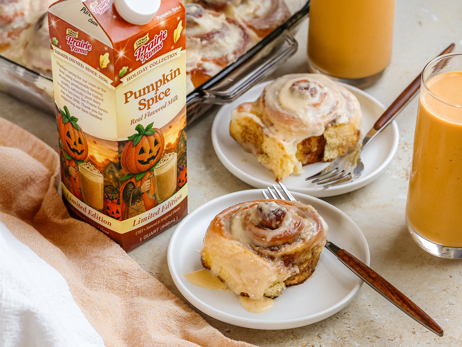 Pumpkin spice cinnamon rolls topped with cream cheese frosting, served on white plates beside glasses of Prairie Farms Pumpkin Spice Milk.