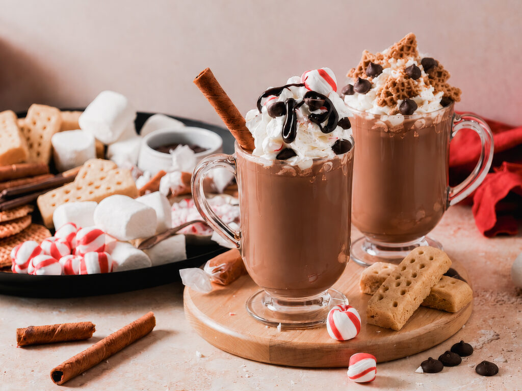 Two mugs of hot chocolate topped with whipped cream and assorted toppings, served beside a board of marshmallows, peppermint candies, cookies, and wafer sticks.