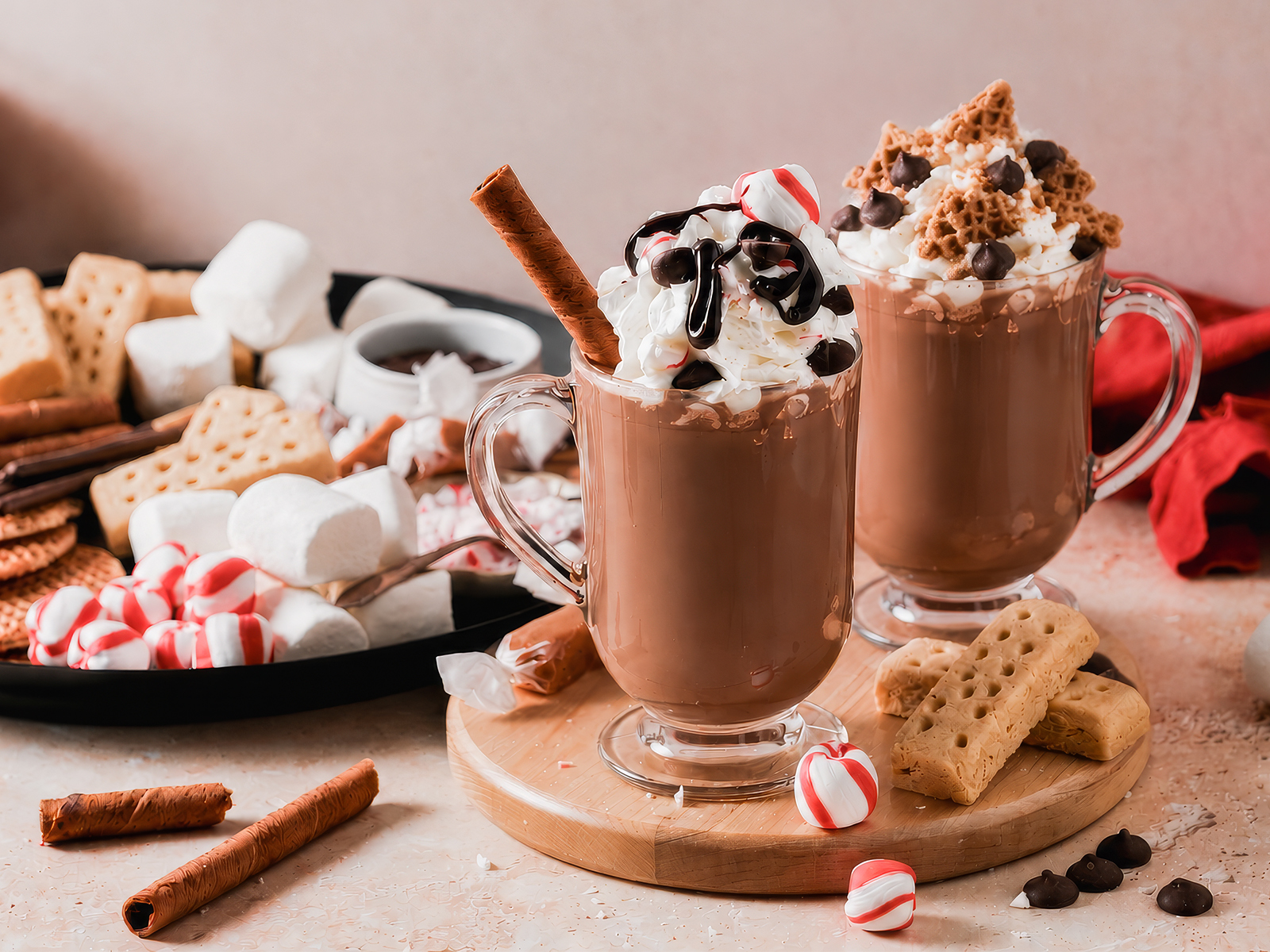 Two mugs of hot chocolate topped with whipped cream and assorted toppings, served beside a board of marshmallows, peppermint candies, cookies, and wafer sticks.