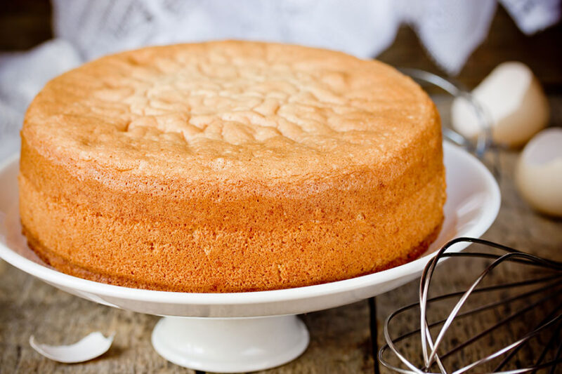 Plain golden sponge cake on a white cake stand, with a whisk nearby.