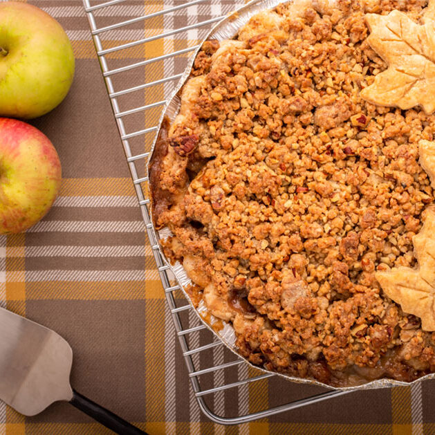 Apple crumb pie with pastry leaves on a cooling rack, beside apples and a pie server