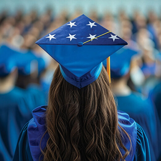 A graduate seen from behind wears a blue cap decorated with white stars while standing among rows of graduates in matching blue gowns at a commencement ceremony