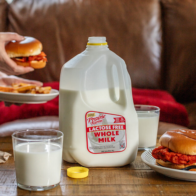Prairie Farms Lactose Free Whole Milk gallon with two glasses of milk and chicken sandwiches on a dining table.