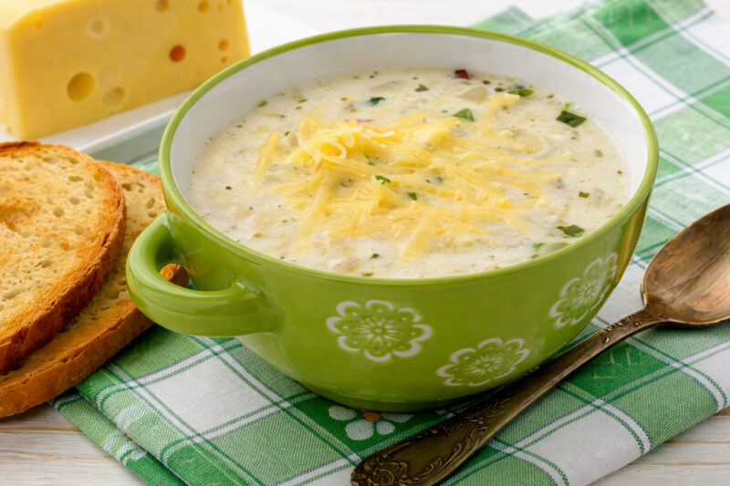 Bowl of creamy potato soup topped with shredded cheese, with bread slices and a spoon beside it.
