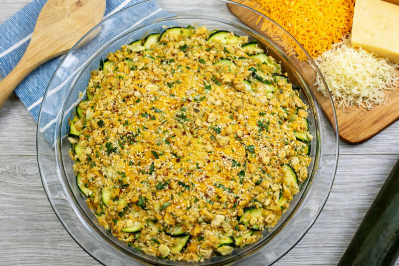 Baking dish with sliced zucchini gratin topped with breadcrumbs and sesame seeds, surrounded by cheese and a wooden spoon