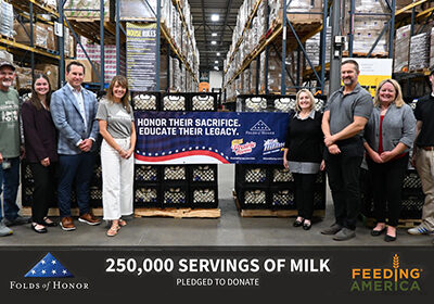 Group of representatives from Folds of Honor, Prairie Farms, Hiland Dairy, and Feeding America standing in a warehouse beside pallets of milk crates and a banner announcing a pledge to donate 250,000 servings of milk to support food banks and military families.