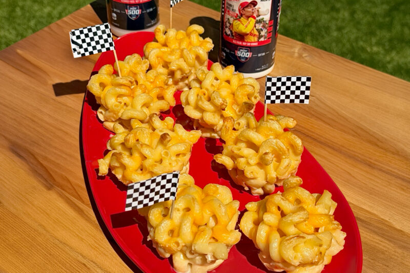 Baked mac and cheese cups arranged on a red platter with checkered racing flags, featuring golden crispy edges and creamy cheesy pasta, with Prairie Farms Milk bottles in the background.