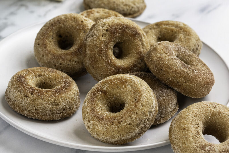 A plate of homemade buttermilk apple cider donuts coated in a cinnamon sugar mixture, stacked closely together on a white dish.