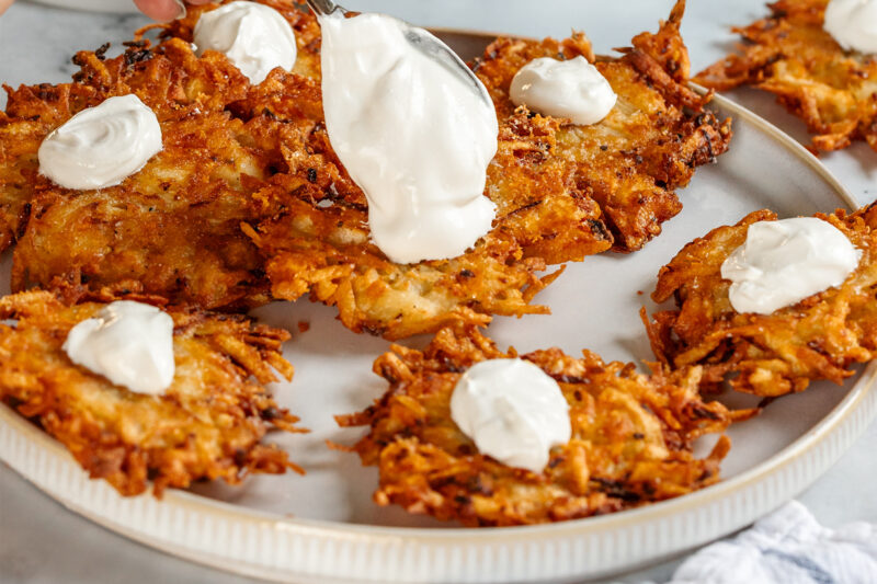Close-up of crispy cheesy latkes on a plate, each topped with sour cream, as a spoon adds a generous dollop to one latke, highlighting the golden, crunchy texture.