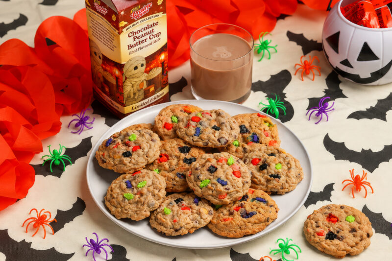 Chocolate peanut butter monster cookies filled with chocolate chips and colorful Halloween sprinkles, served with a glass of Prairie Farms Chocolate Peanut Butter Cup Milk among festive bats and spiders.