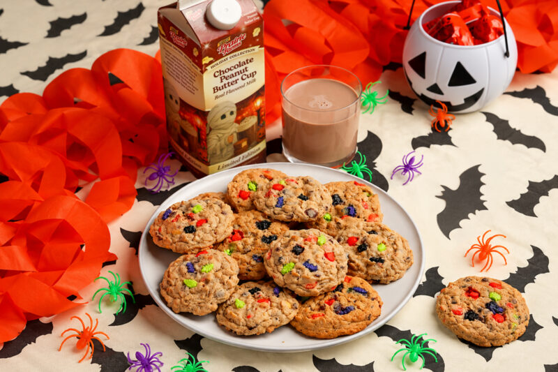 Chocolate peanut butter monster cookies filled with chocolate chips and colorful Halloween sprinkles, served with a glass of Prairie Farms Chocolate Peanut Butter Cup Milk among festive bats and spiders.