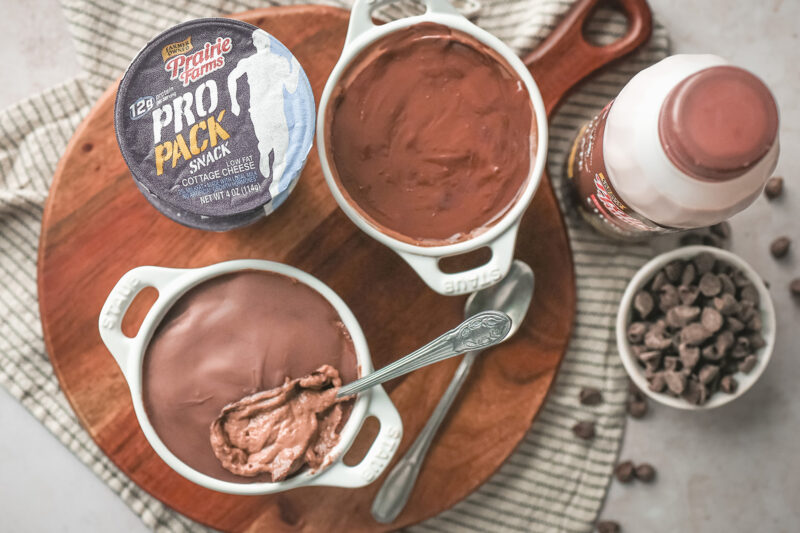 Overhead view of two chocolate protein cheesecake cups in small baking dishes on top of a circular wooden serving board, with one cracked open showing the creamy texture.