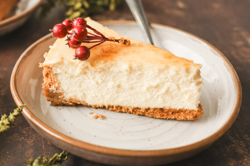 A creamy slice of crème brûlée cheesecake with a golden top, garnished with festive red berries and served on a ceramic plate.