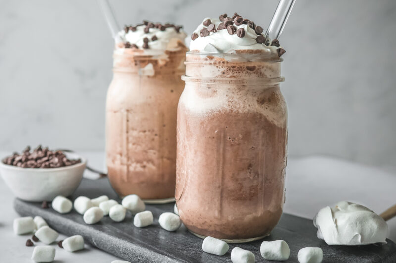 Two mason jars filled with frozen mint hot chocolate topped with whipped cream and mini chocolate chips, served with glass straws and surrounded by mini marshmallows on a dark slate board.