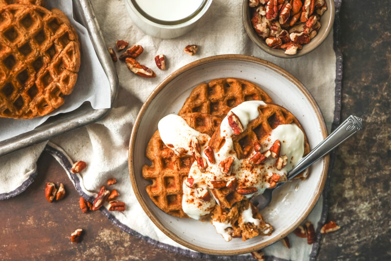Plate of golden gingerbread waffles topped with creamy drizzle and chopped pecans, served with a glass of milk nearby.