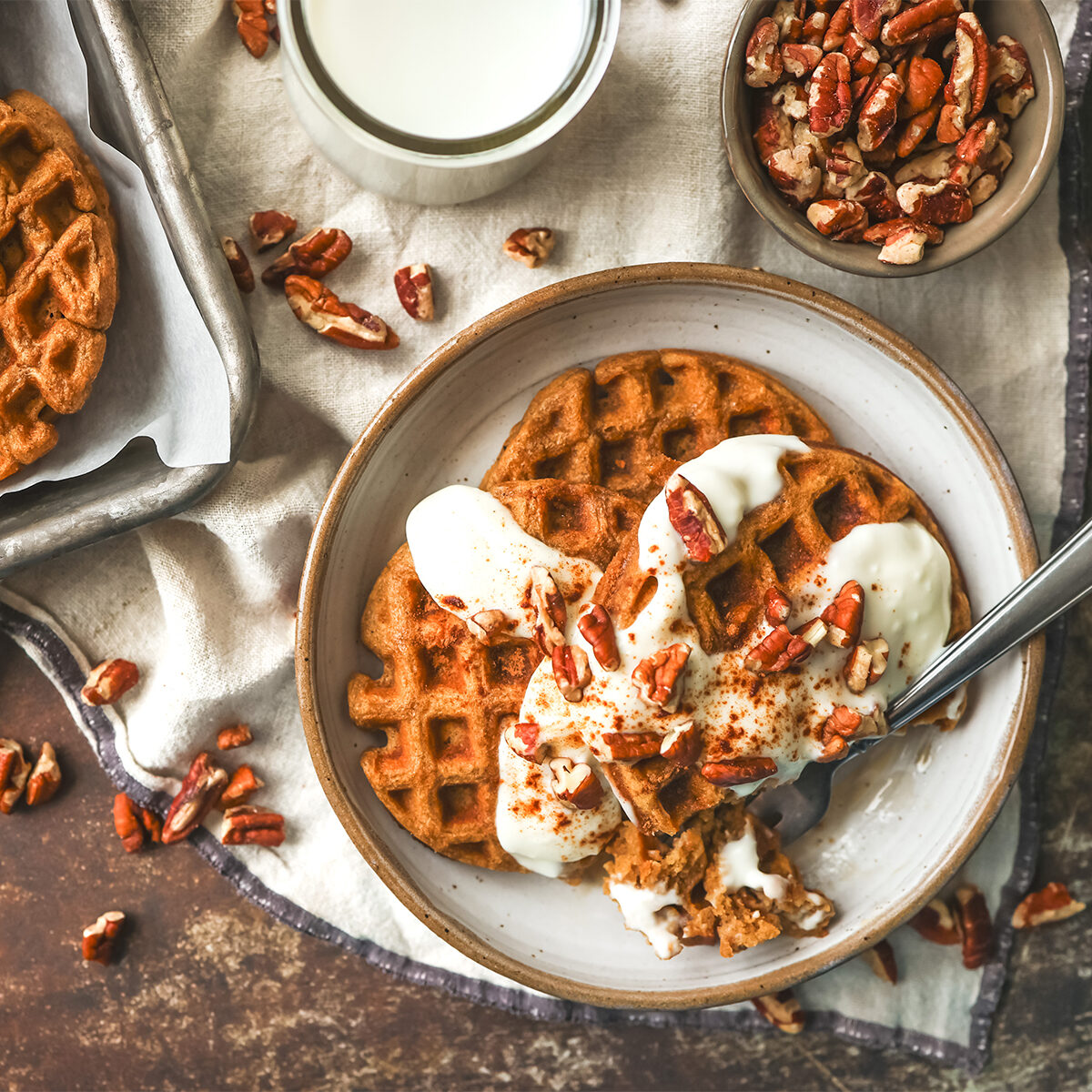 Plate of golden gingerbread waffles topped with creamy drizzle and chopped pecans, served with a glass of milk nearby.