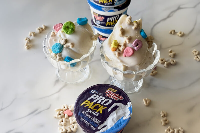 Two dessert bowls filled with creamy cottage cheese ice cream topped with marshmallow cereal pieces, with cottage cheese snack cups stacked in the background and cereal scattered on the surface.