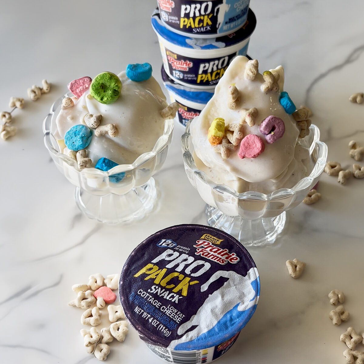 Two dessert bowls filled with creamy cottage cheese ice cream topped with marshmallow cereal pieces, with cottage cheese snack cups stacked in the background and cereal scattered on the surface.