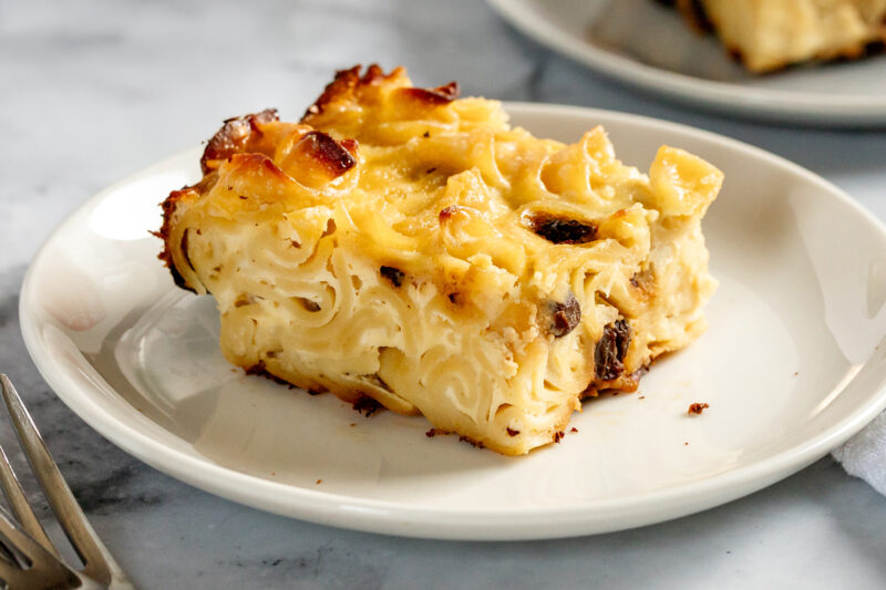 A slice of baked noodle kugel on a white plate, showing tender noodles with a lightly golden, caramelized top and creamy interior.