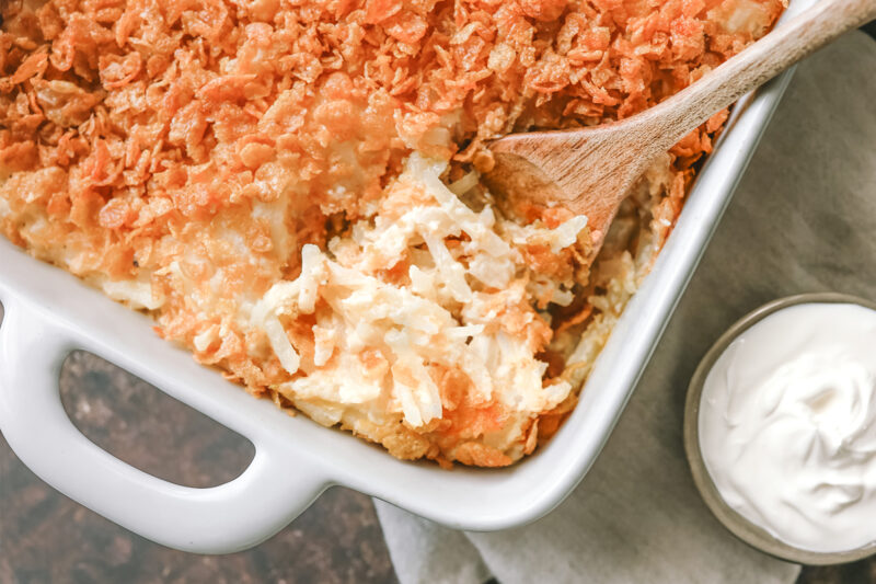 Creamy hash brown casserole topped with a crispy cornflake crust, served with a side of sour cream in a white baking dish.
