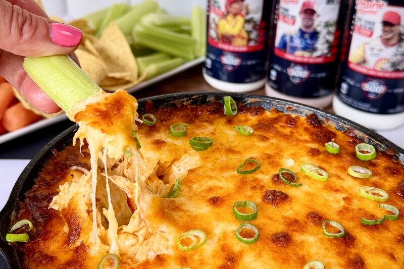 A cheesy baked buffalo chicken skillet dip topped with sliced green onions, as a celery stick lifts a gooey, melted portion, with chips, celery sticks and milk bottles in the background.
