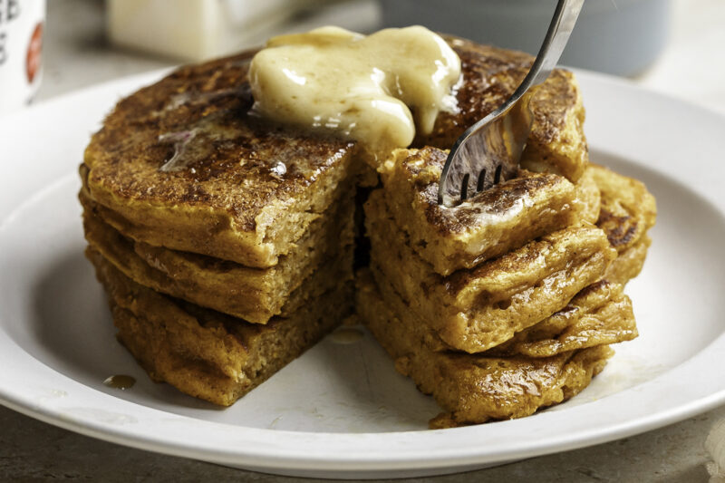 Stack of fluffy pumpkin cottage cheese pancakes on a white plate, topped with melting butter and syrup, with a fork cutting into the stack to reveal the soft, moist texture inside.