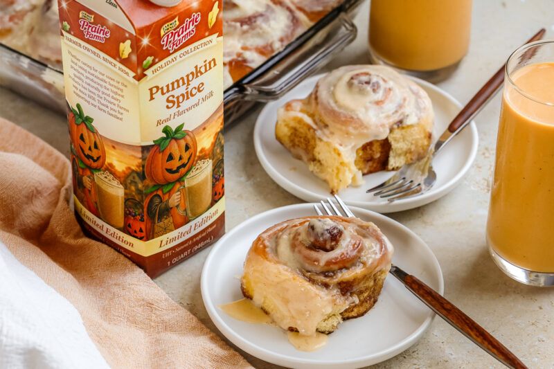 Pumpkin spice cinnamon rolls topped with cream cheese frosting, served on white plates beside glasses of Prairie Farms Pumpkin Spice Milk.