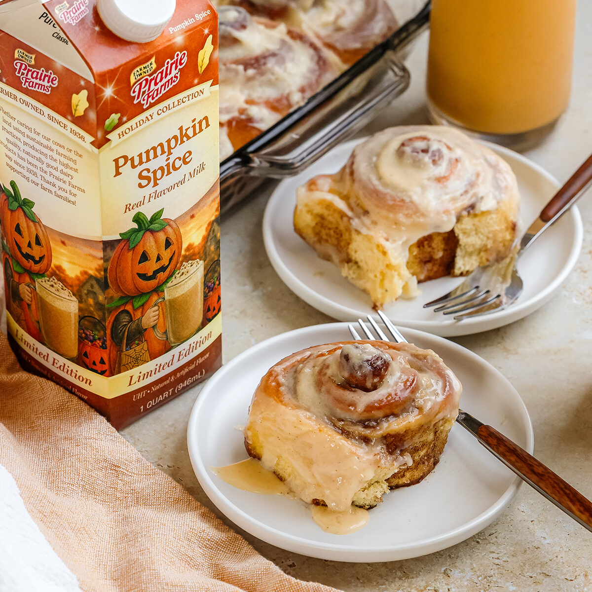 Pumpkin spice cinnamon rolls topped with cream cheese frosting, served on white plates beside glasses of Prairie Farms Pumpkin Spice Milk.