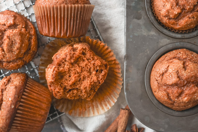 Freshly baked pumpkin spice muffins in a metal muffin tin and on a cooling rack, with one muffin cut open to show its soft, moist texture.