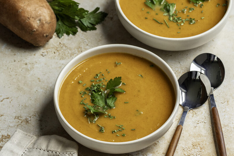 Two bowls of creamy roasted fall vegetable soup topped with fresh parsley, set on a light stone surface with spoons beside them and a sweet potato and parsley in the background.
