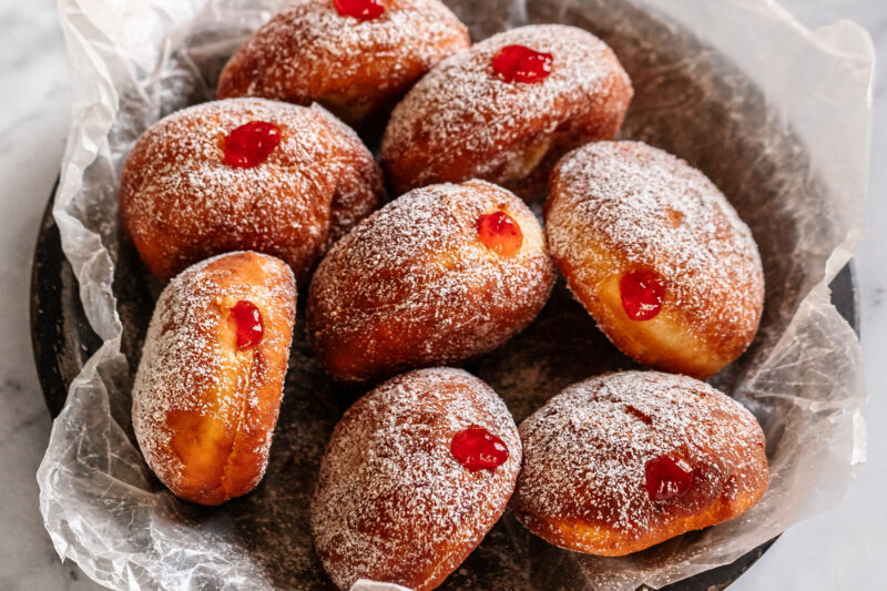 A dark bowl lined with parchment paper containing eight jelly-filled fried doughnuts dusted with powdered sugar, each topped with a bright red jelly center.